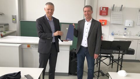 Kultusstaatssekretär Dr. Manuel Lösel übergibt den Bescheid, beide lächeln in die Kamera