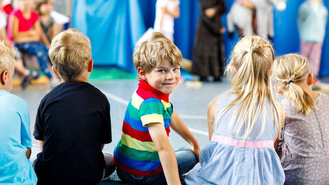 Kindertheater macht Spaß Kinder sitzen in eienr Reihe und gucken einer Theater Aufführung zu. Ein Kind dreht sich um und lacht in die Kamera.
