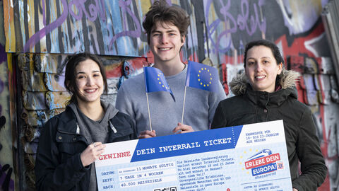 Drei junge Menschen stehen vor einer Graffiti besprühten Wand mit einem "HessenRail"-Scheck in Händen. Die mittlere Person wedelt mit kleinen Europa-Flaggen