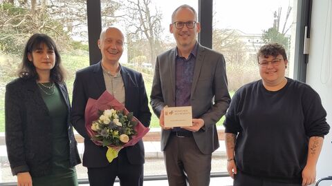 Sozialminister Kai Klose und Knud Wechterstein stehen mit zwei weiteren Personen vor Fenster. Kai Klose hält Urkunde in der Hand.