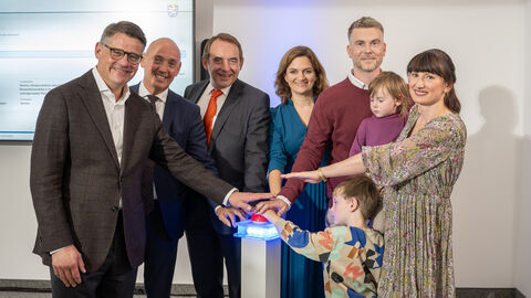 V. l. n. r.: Ministerpräsident Boris Rhein, WIBank-Sprecher Gottfried Milde, Finanzminister Alexander Lorz, WIBank-Geschäftsführerin Judith Mandel und die Hessengeld-Empfänger-Familie, Tobias und Isabel Zöller mit ihren Kindern.  Sechs Erwachsende und zwei Kinder stehen in einem Halbkreis und strecken jeweils einen Arm in die Mitte.